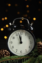 Photo of Alarm clock, fir tree branches and cones on table against blurred festive lights