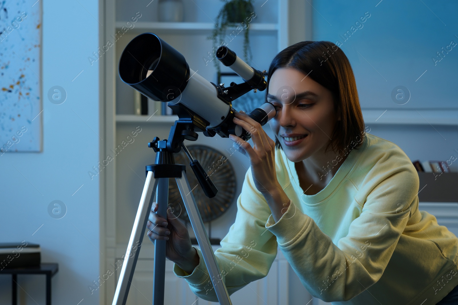 Photo of Beautiful young woman looking at stars through telescope in room