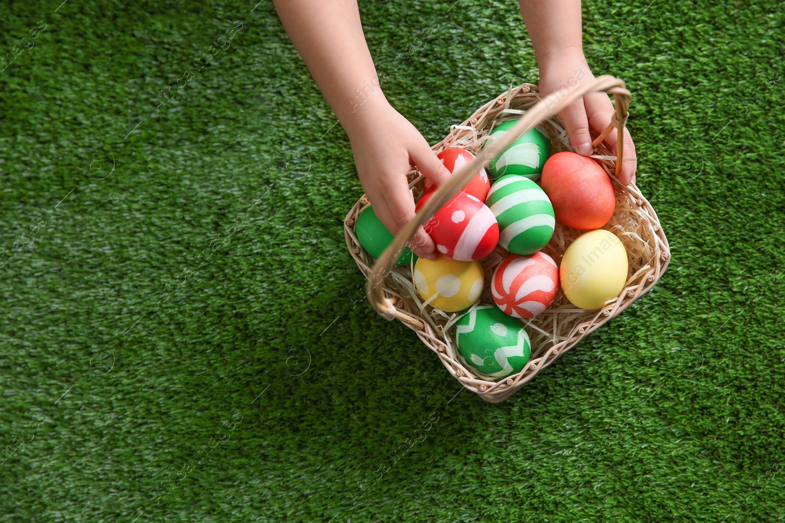 Photo of Little child with basket of painted Easter eggs on green grass, top view. Space for text