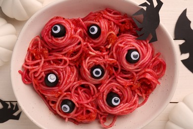 Red pasta with decorative eyes and olives in bowl served on white wooden table, flat lay. Halloween food
