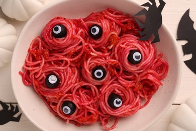 Photo of Red pasta with decorative eyes and olives in bowl served on white wooden table, flat lay. Halloween food