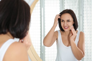 Photo of Happy mature woman cleaning face with cotton pad near mirror in bathroom