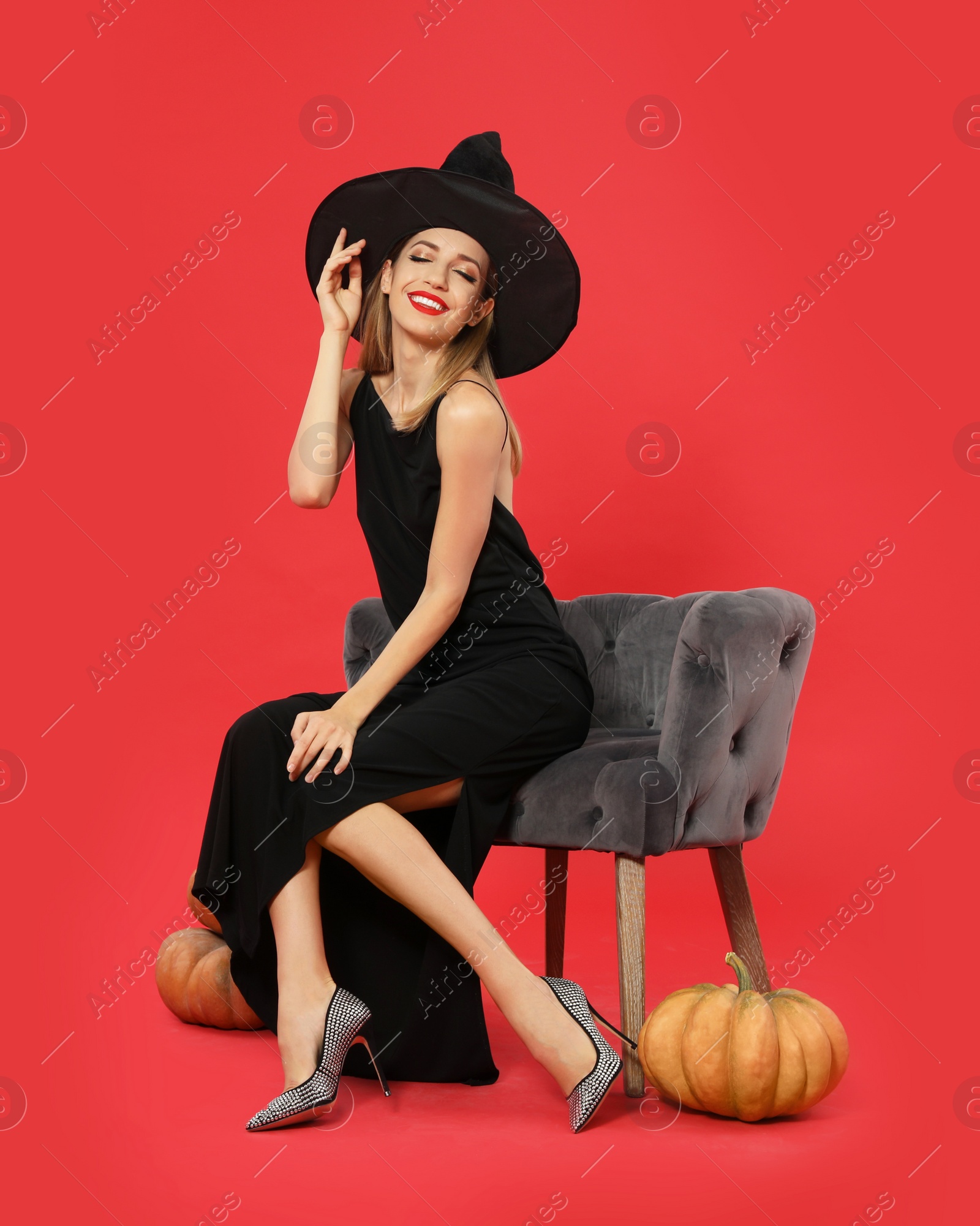 Photo of Beautiful woman wearing witch costume in armchair and pumpkins on red background, space for text. Halloween party