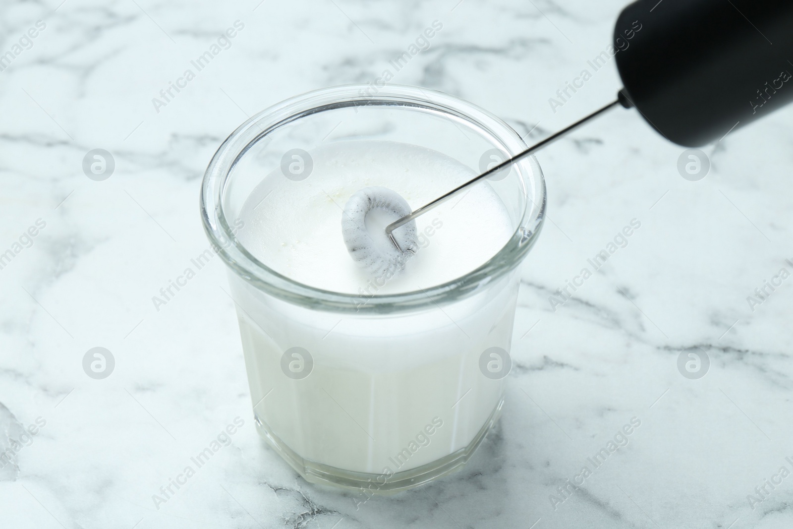 Photo of Whisking milk in glass with mini mixer (milk frother) at white marble table, closeup