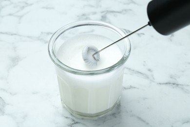 Whisking milk in glass with mini mixer (milk frother) at white marble table, closeup