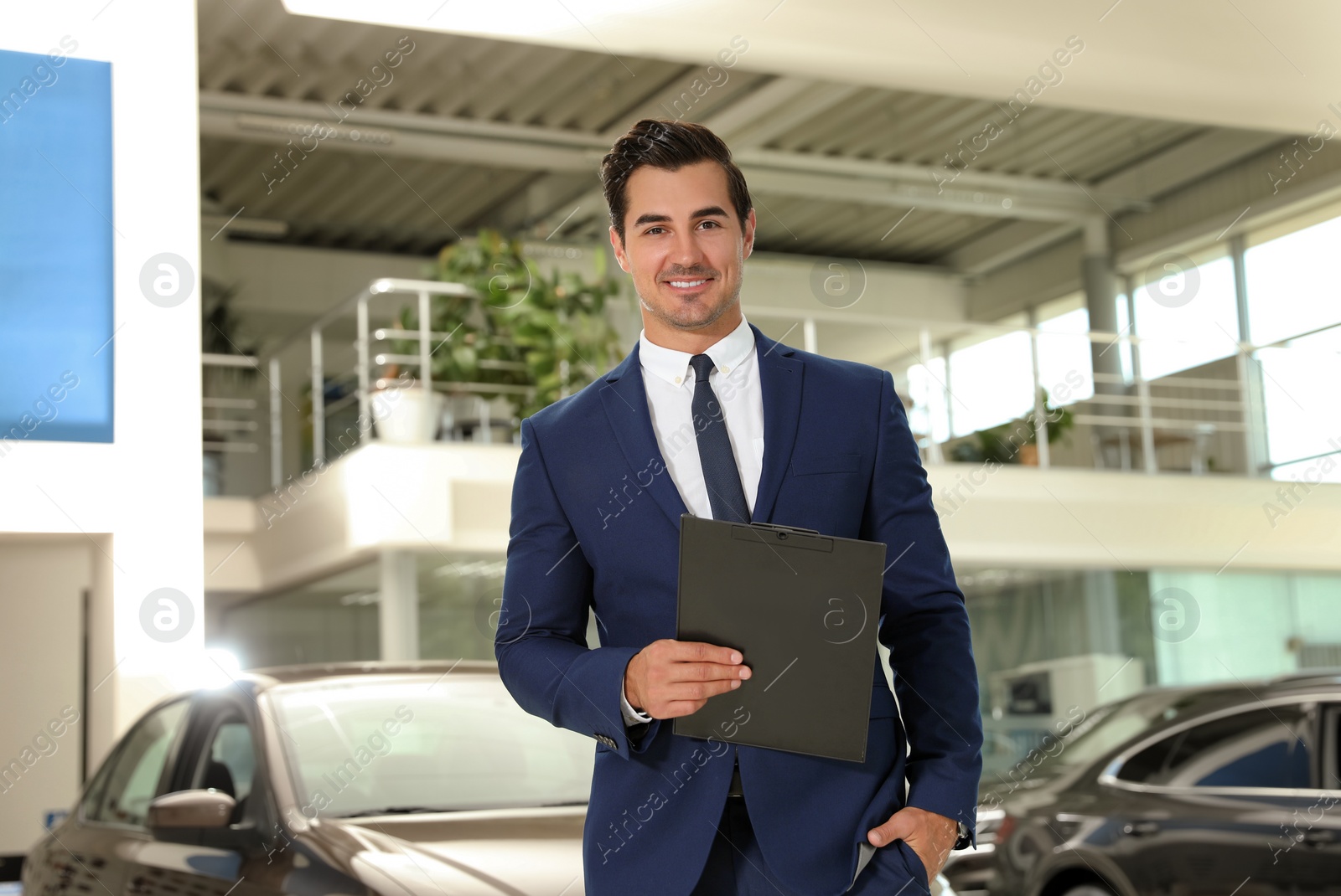 Photo of Young salesman with clipboard near car in modern dealership