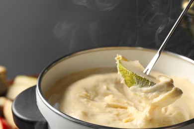 Photo of Dipping piece of pear into fondue pot with melted cheese on grey background, closeup