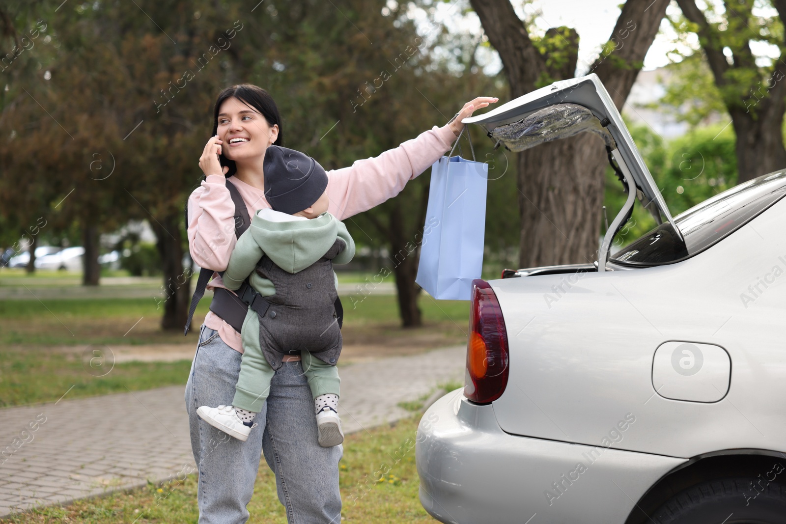 Photo of Mother holding her child in sling (baby carrier) while closing car trunk outdoors