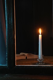 Photo of Burning candle and Bible on wooden table at night, view through window
