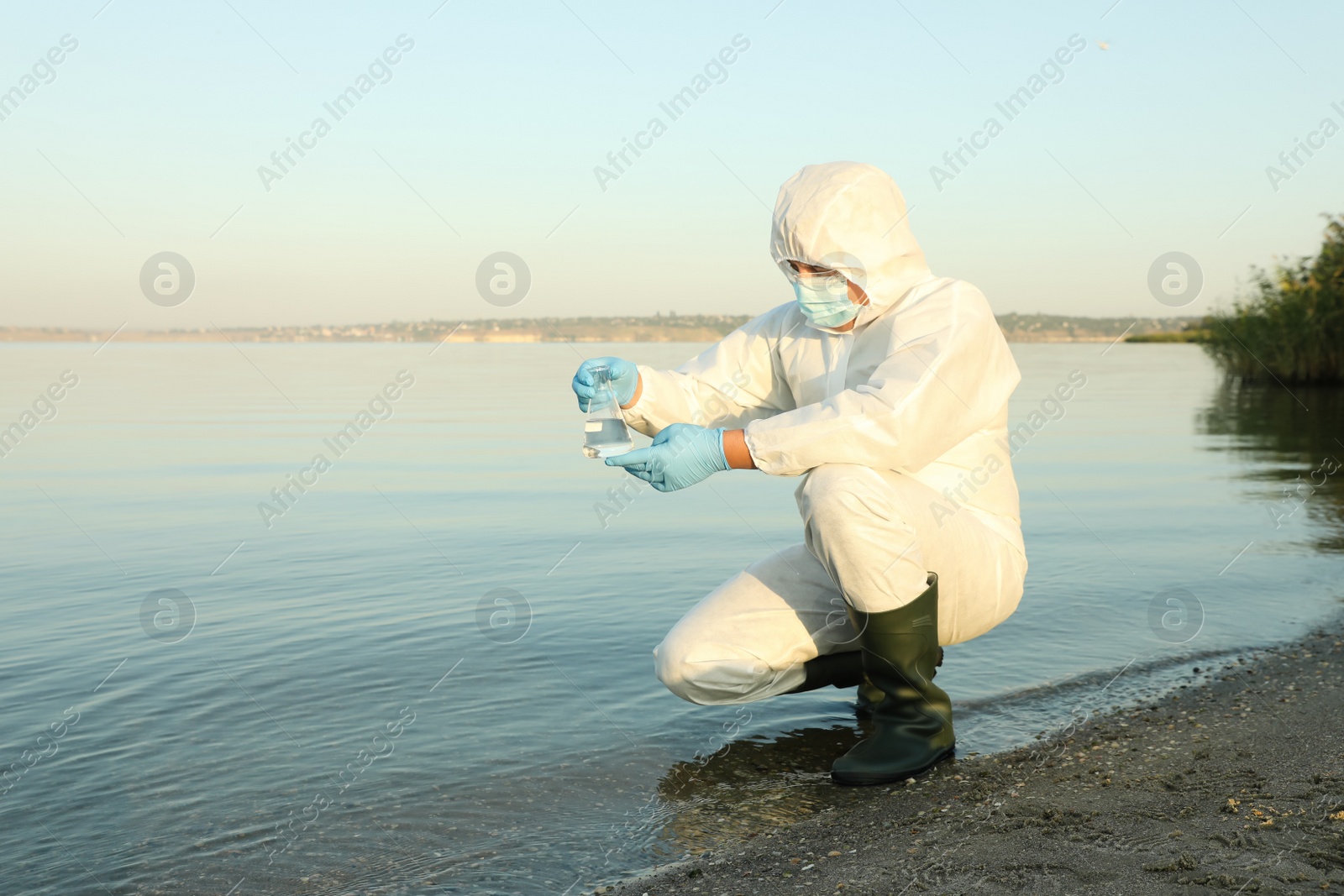 Photo of Scientist in chemical protective suit with conical flask taking sample from river for analysis