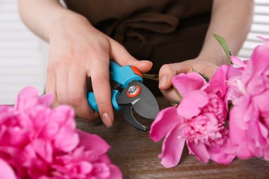 Photo of Woman trimming beautiful pink peonies with secateurs at wooden table, closeup