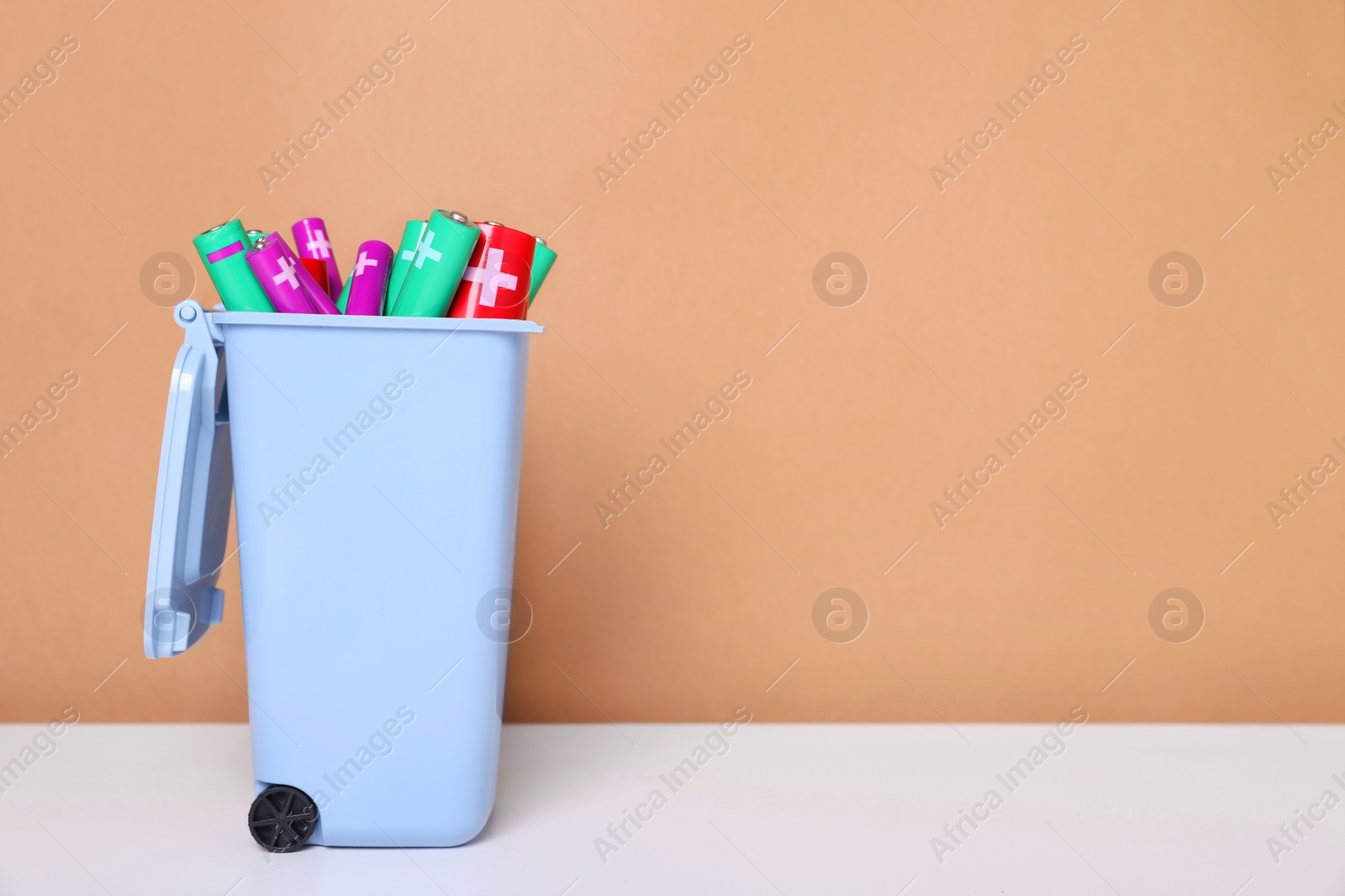 Photo of Many used batteries in recycling bin on white table against light brown background. Space for text