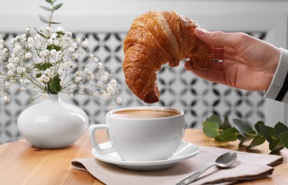 Tasty break. Woman dipping fresh croissant into cup with cappuccino at wooden table indoors, closeup