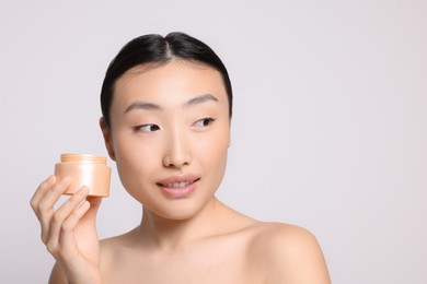 Photo of Beautiful young Asian woman holding jar of body cream on light grey background. Space for text