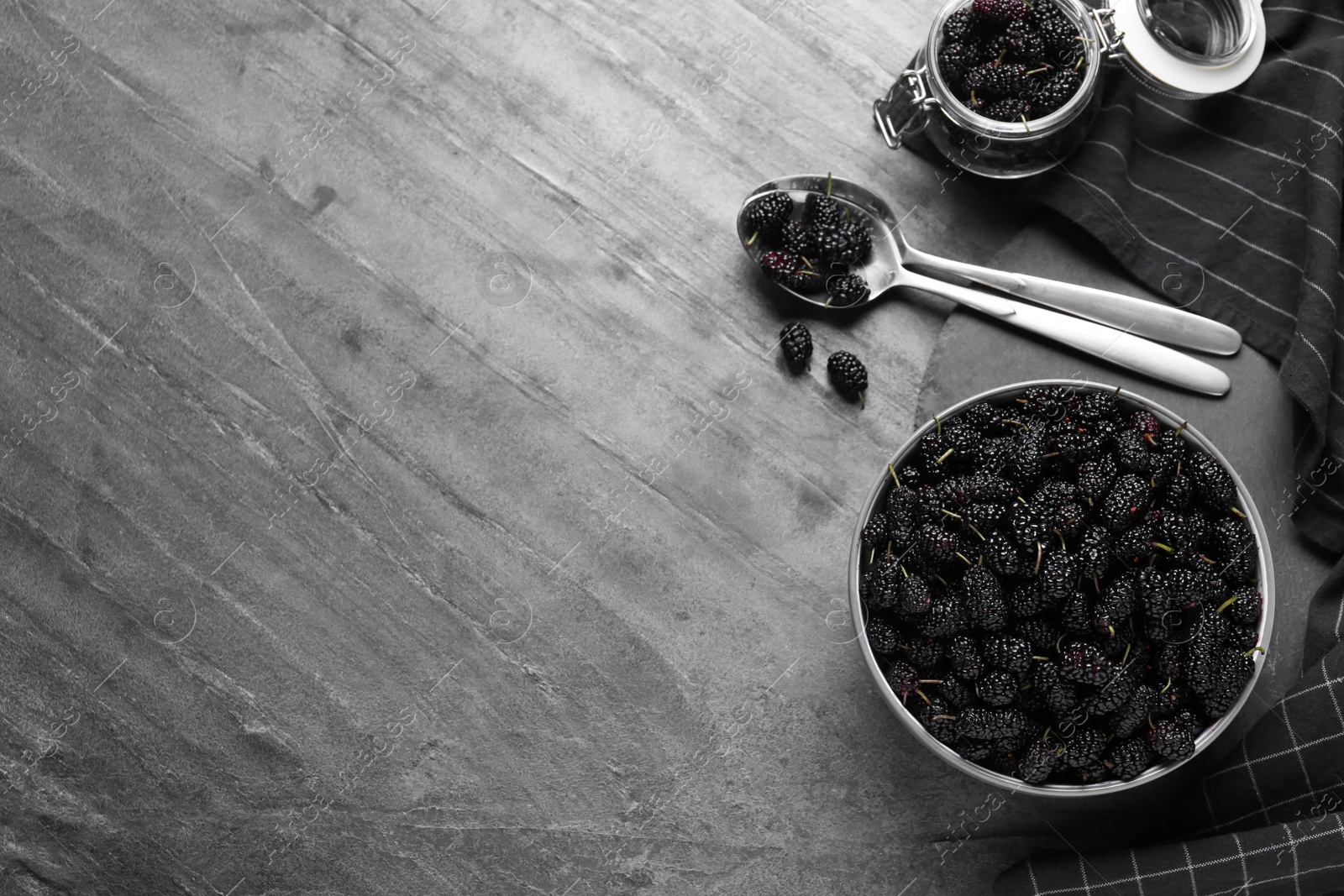 Photo of Bowl, spoon and jar of delicious ripe black mulberries on grey table, flat lay. Space for text