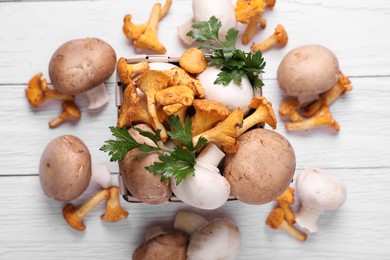 Basket with different mushrooms and parsley on white wooden table, flat lay