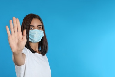 Photo of Young woman in protective mask showing stop gesture on light blue background, space for text. Prevent spreading of coronavirus