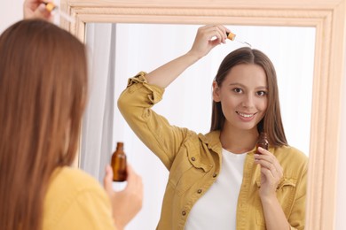 Photo of Beautiful woman applying serum onto hair near mirror indoors