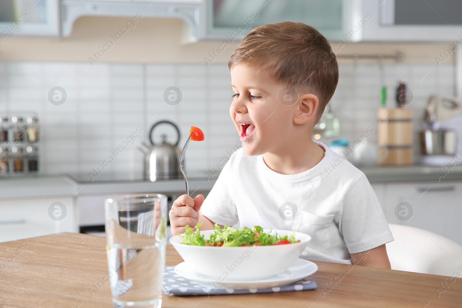 Photo of Adorable little boy eating vegetable salad at table in kitchen