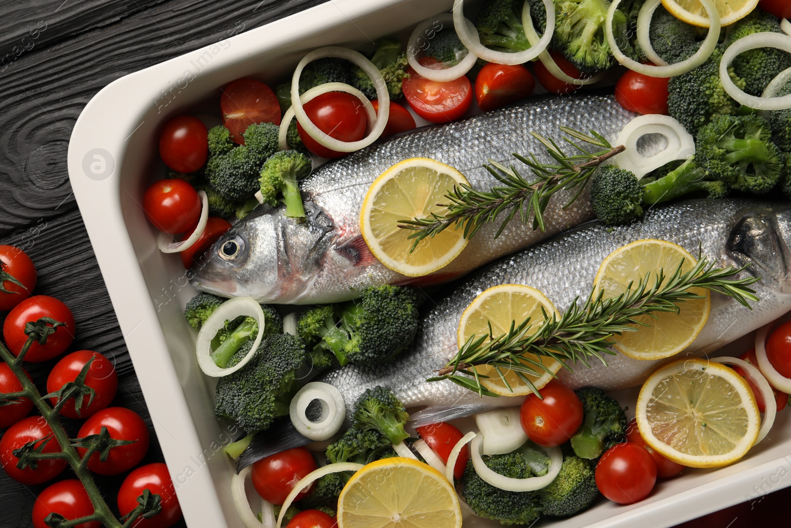 Photo of Raw fish with lemon in baking dish and vegetables on black wooden table, flat lay