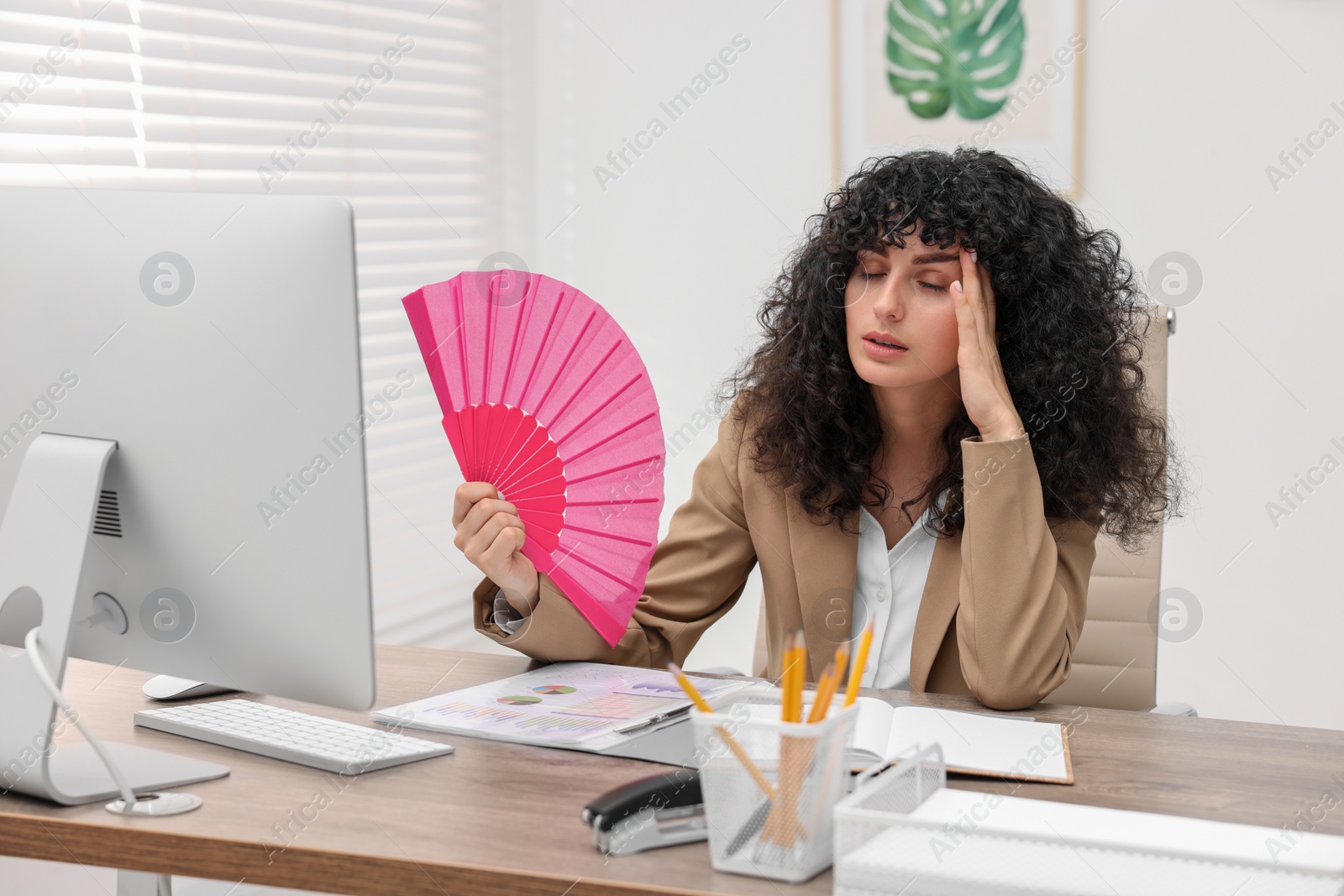 Photo of Young businesswoman waving hand fan to cool herself at table in office