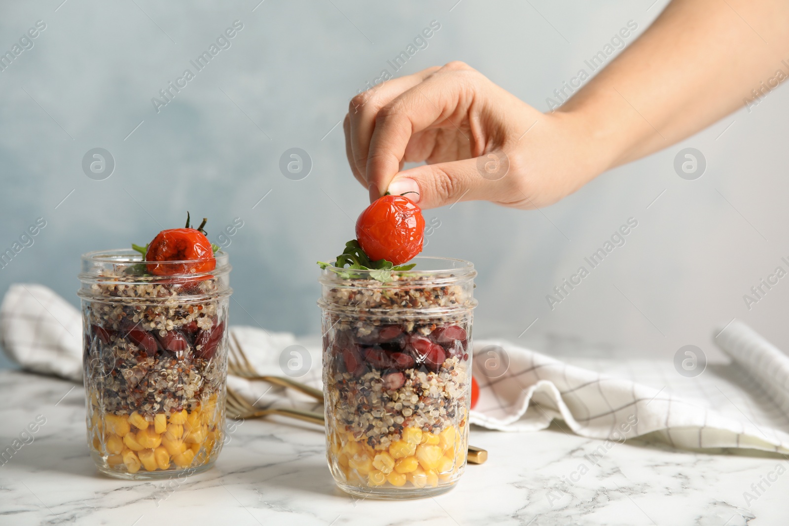 Photo of Woman putting tomato into jar with healthy quinoa salad and vegetables on table, closeup