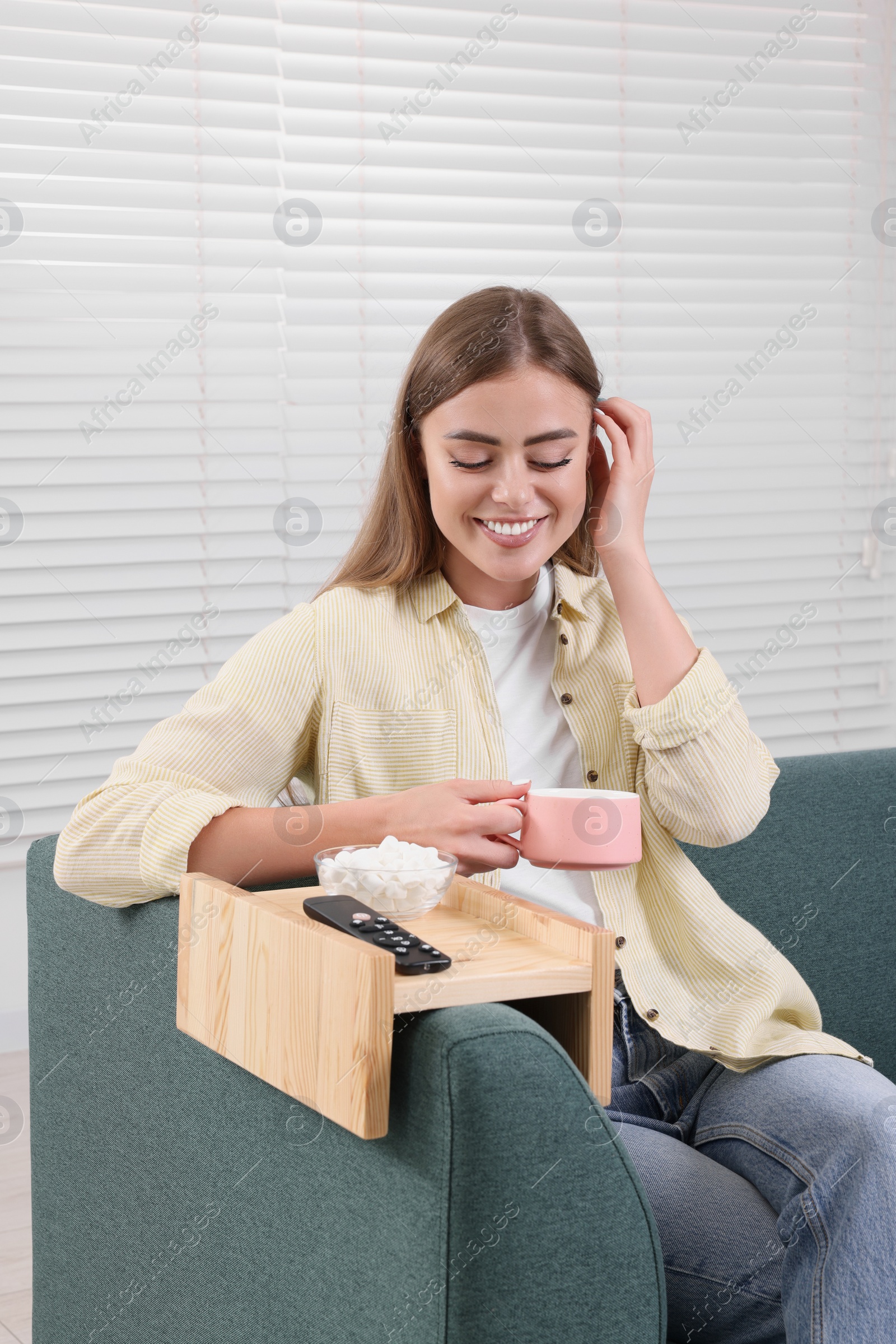 Photo of Happy woman holding cup of drink at home. Marshmallows and remote control on sofa armrest wooden table