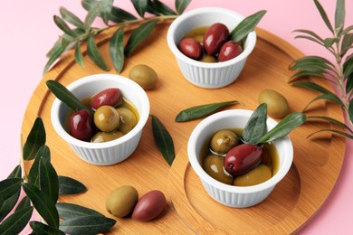 Wooden tray with different ripe olives and leaves on pink background