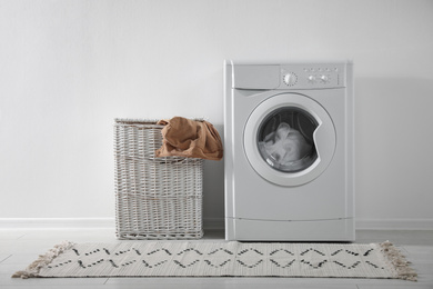 Photo of Modern washing machine and laundry basket near white wall indoors. Bathroom interior