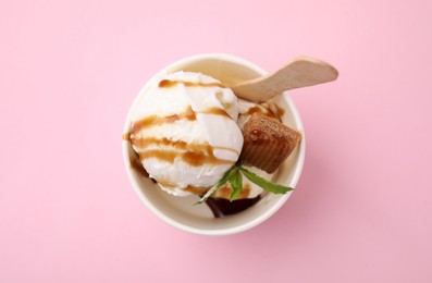 Tasty ice cream with caramel sauce, mint leaves and candy in paper cup on pink background, top view