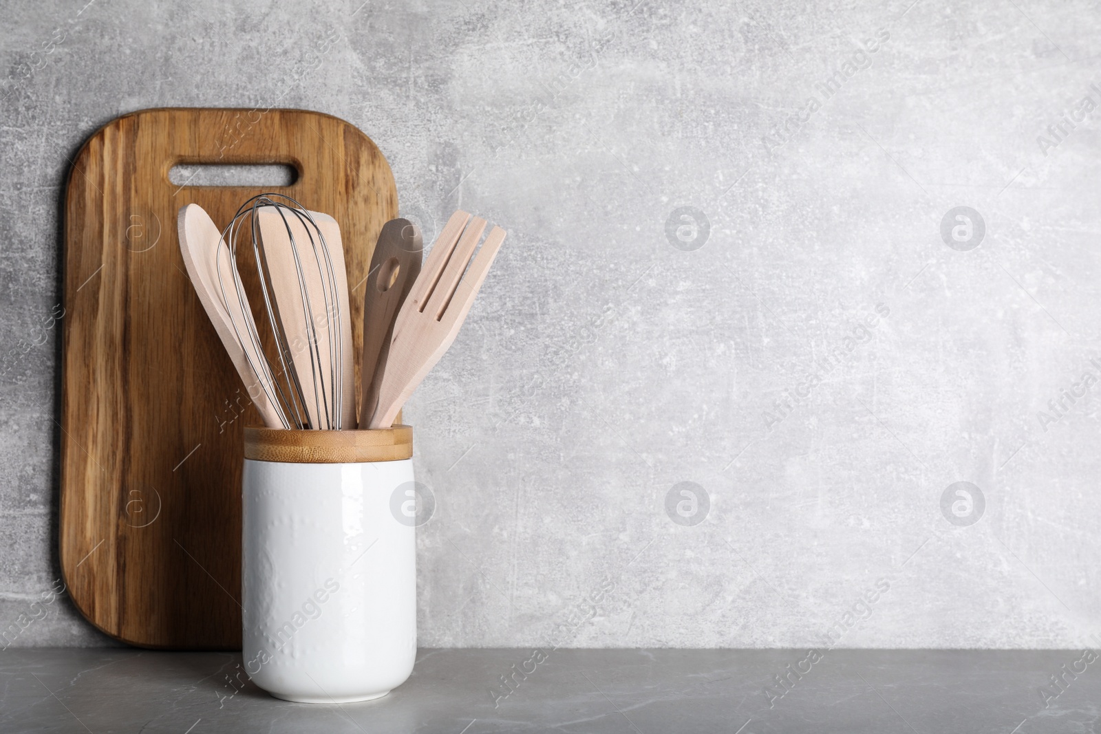 Photo of Holder with wooden kitchen utensils and cutting board on grey table. Space for text