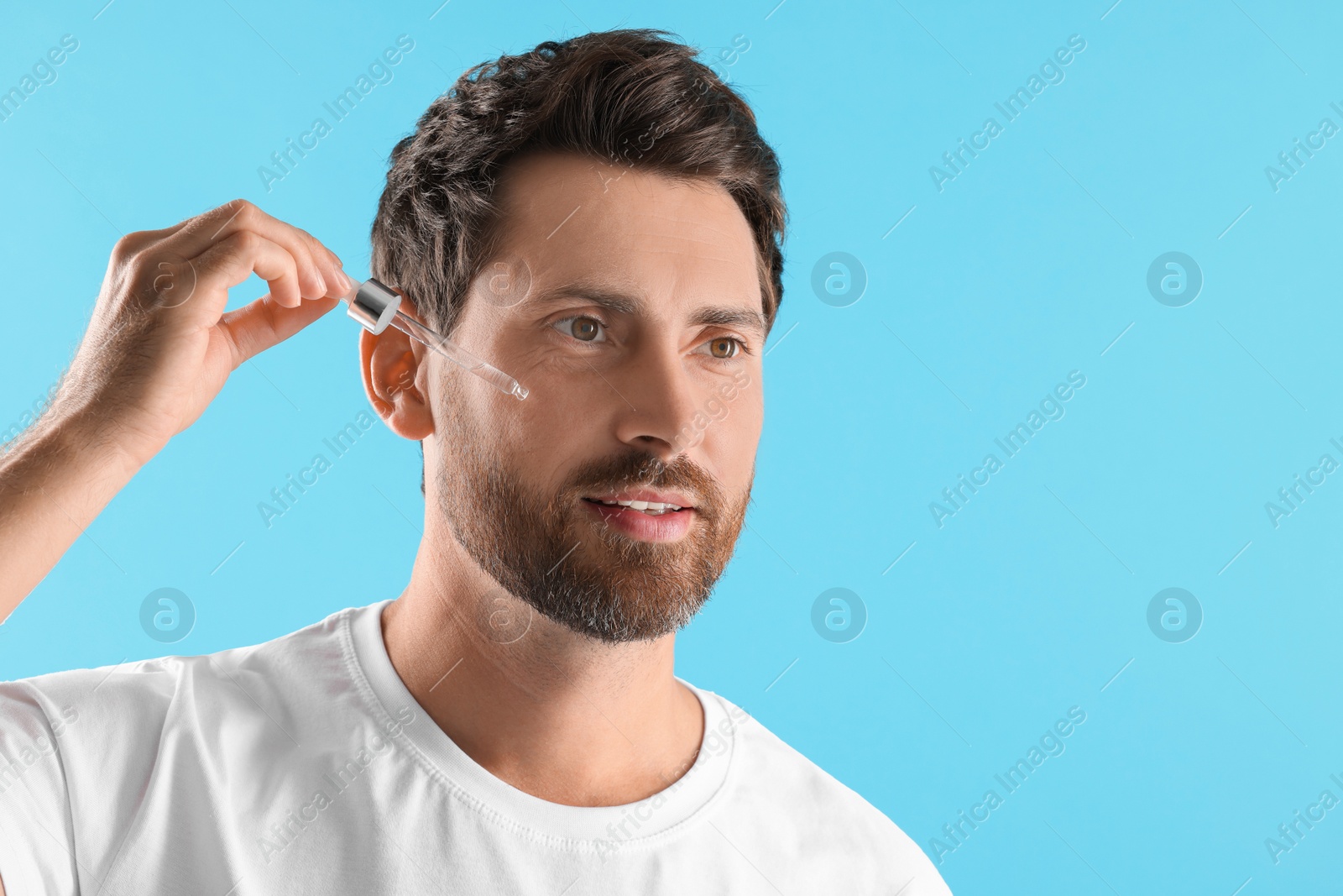 Photo of Handsome man applying cosmetic serum onto his face on light blue background. Space for text