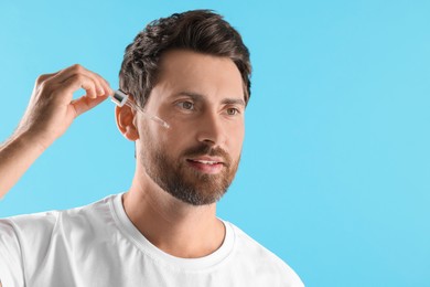 Handsome man applying cosmetic serum onto his face on light blue background. Space for text