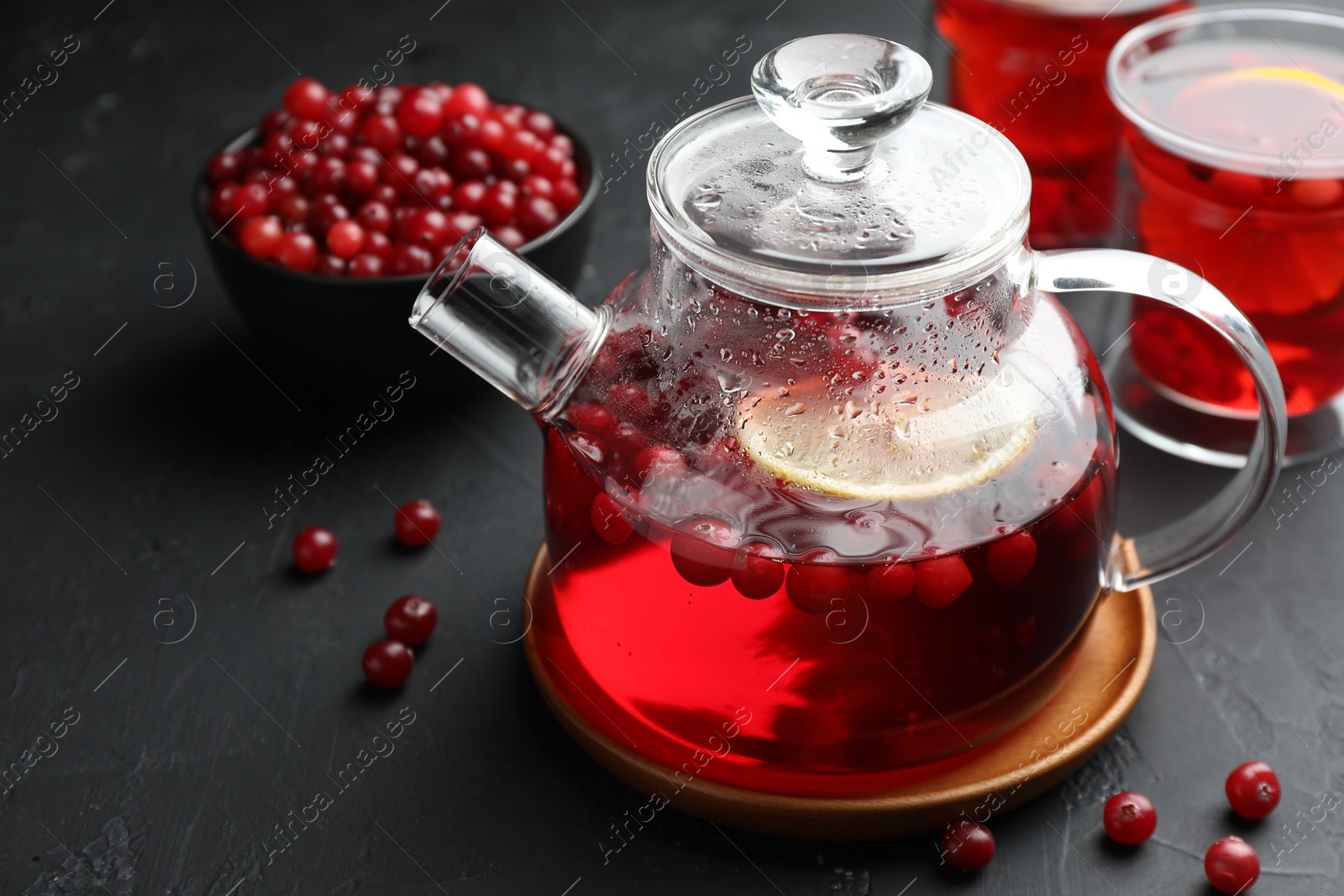 Photo of Tasty hot cranberry tea with lemon in teapot and fresh berries on black textured table