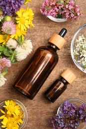 Bottles of essential oils surrounded by beautiful flowers on wooden table, flat lay