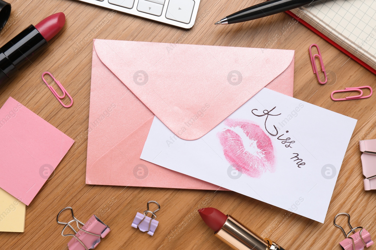 Photo of Card with phrase Kiss me, lipstick mark and envelope on wooden office table, flat lay