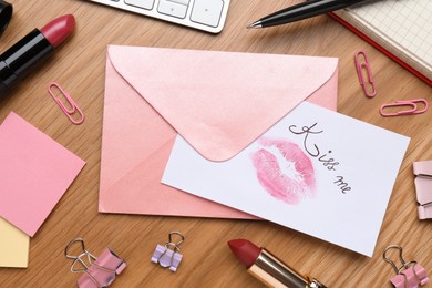Photo of Card with phrase Kiss me, lipstick mark and envelope on wooden office table, flat lay