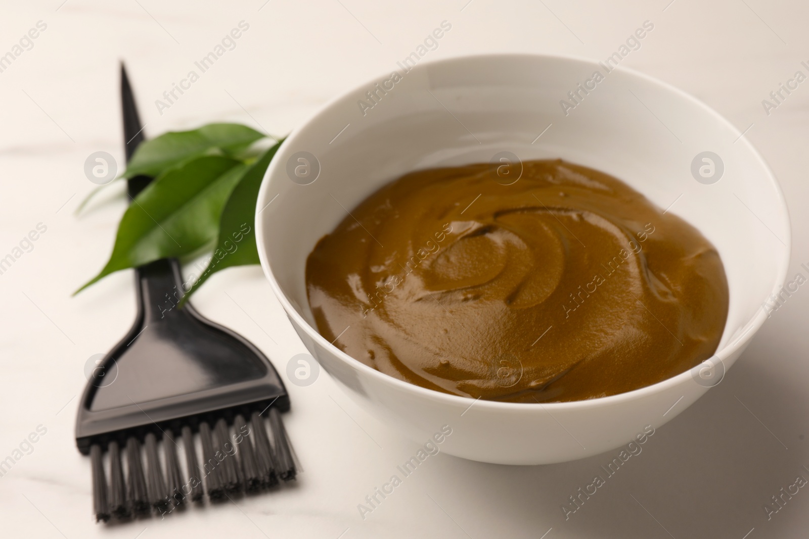 Photo of Bowl of henna cream and brush on white marble table, closeup. Natural hair coloring