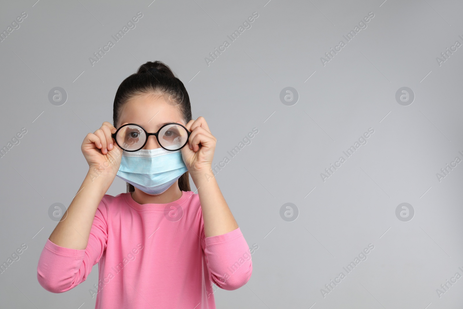 Photo of Little girl wiping foggy glasses caused by wearing medical face mask on grey background, space for text. Protective measure during coronavirus pandemic