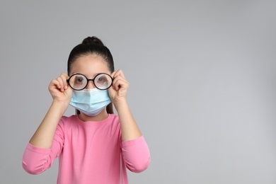 Little girl wiping foggy glasses caused by wearing medical face mask on grey background, space for text. Protective measure during coronavirus pandemic