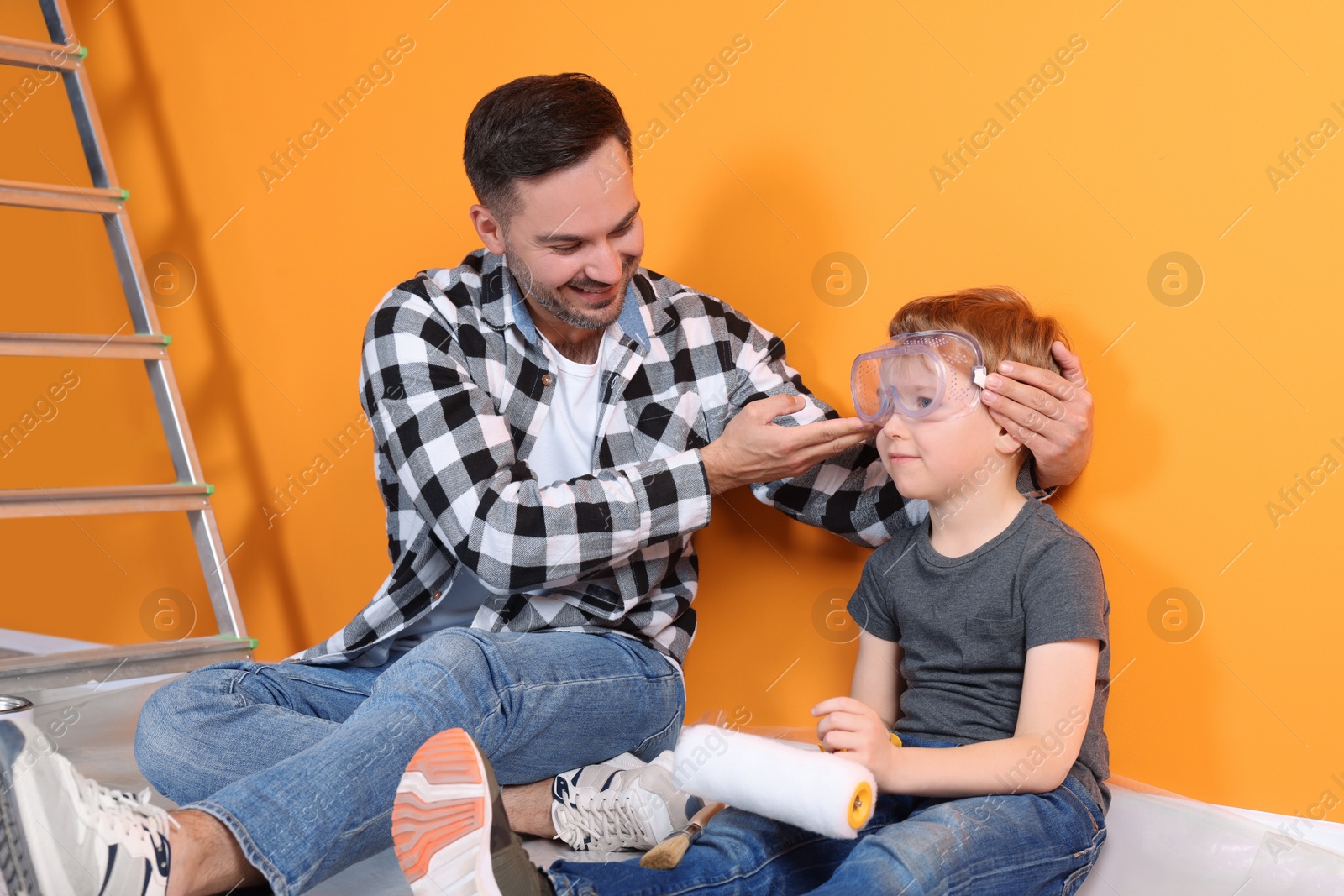 Photo of Father putting protective glasses on his son near orange wall. Repair work