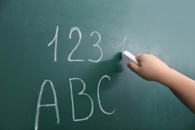 Photo of Little child writing letters and numbers on chalkboard, closeup