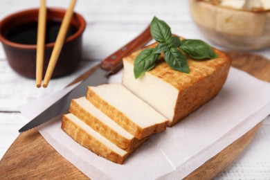 Photo of Board with smoked tofu, knife, basil and soy sauce on white wooden table