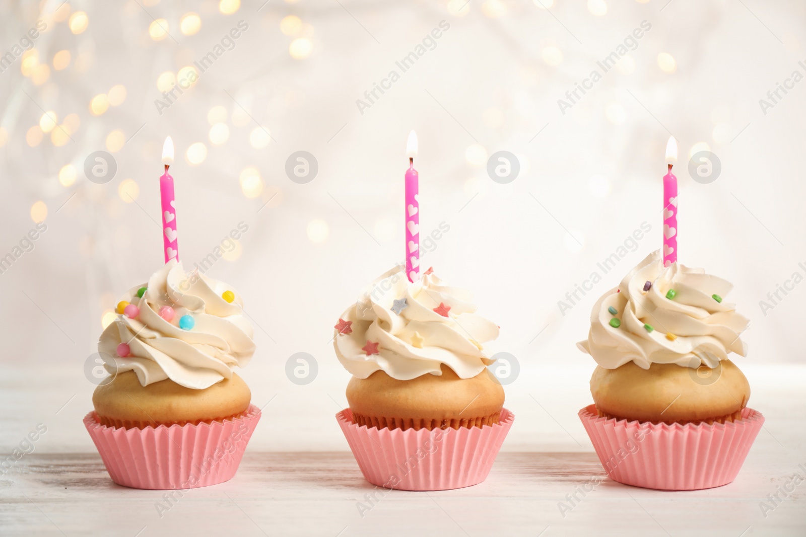 Photo of Birthday cupcakes with candles on white wooden table against blurred lights