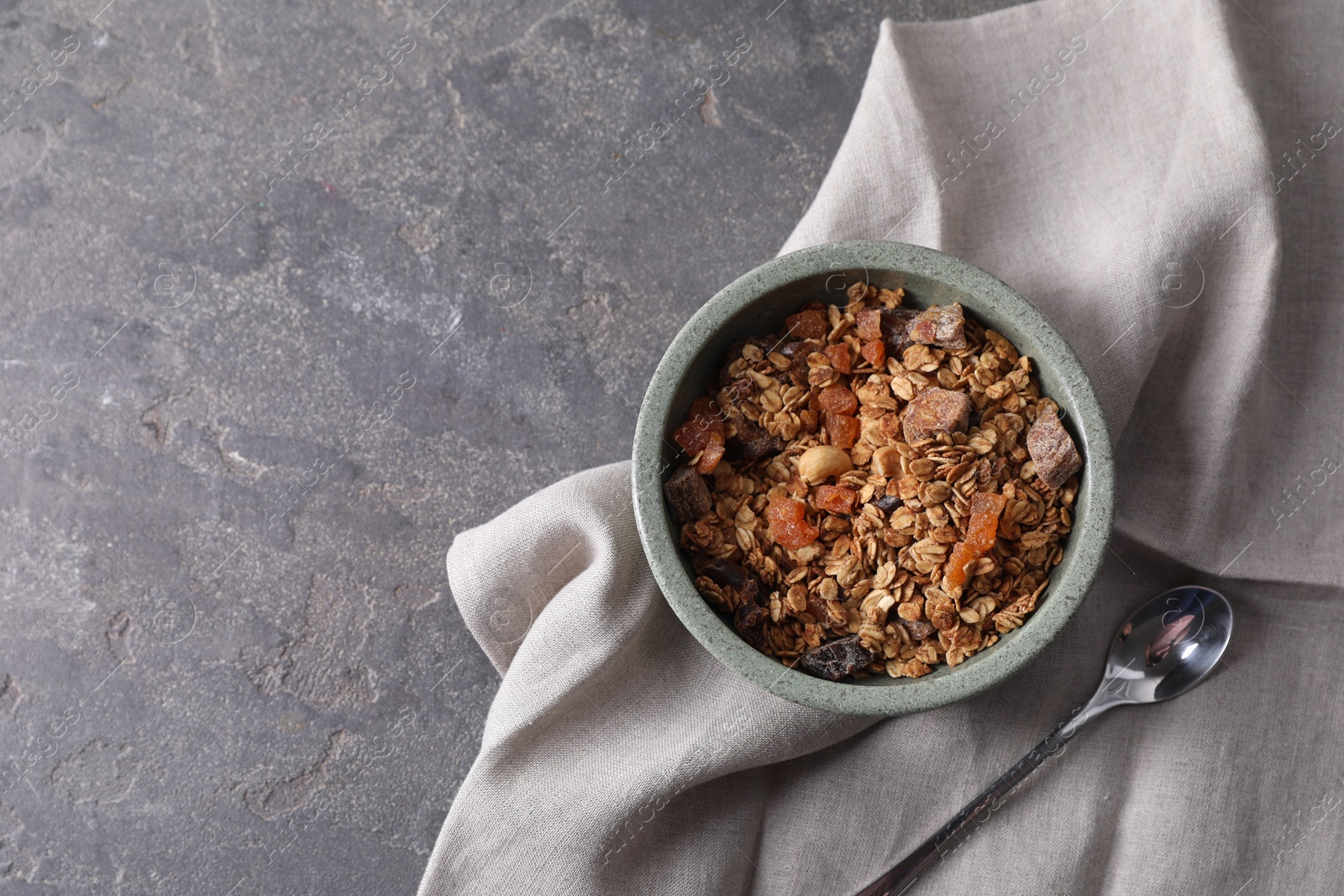 Photo of Tasty granola in bowl, spoon and napkin on gray textured table, flat lay. Space for text