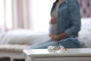 Photo of Cigarettes in ash tray and blurred pregnant woman on background, closeup. Space for text