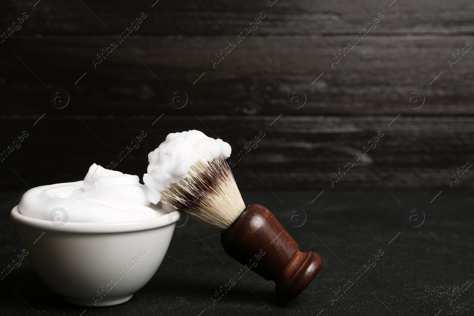 Photo of Shaving brush and bowl of foam on table against dark background with space for text