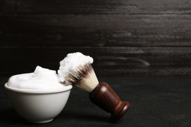 Photo of Shaving brush and bowl of foam on table against dark background with space for text