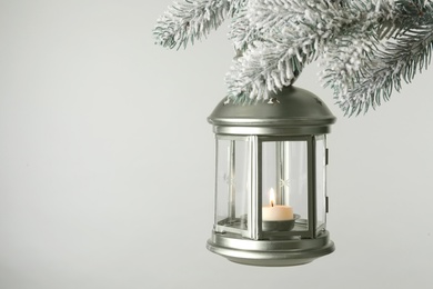 Photo of Christmas lantern with candle hanging on snowy fir tree branch against light background. Space for text