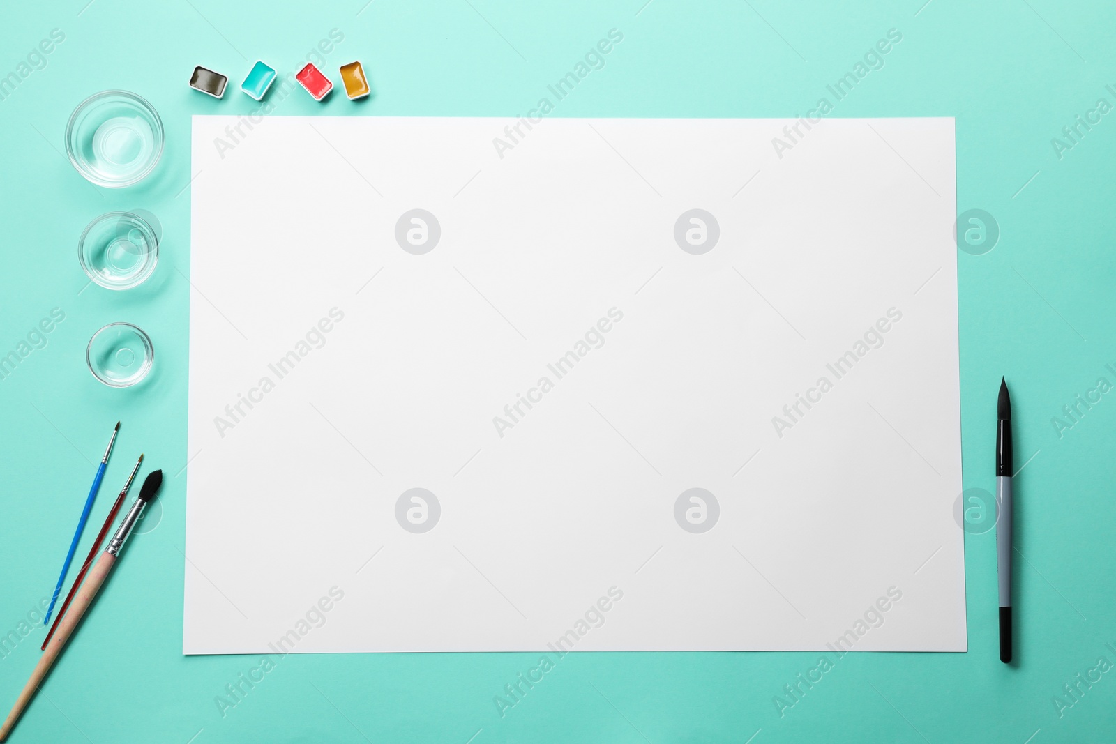 Photo of Blank sheet of paper, watercolor paints, empty bowls and brushes on turquoise background, flat lay